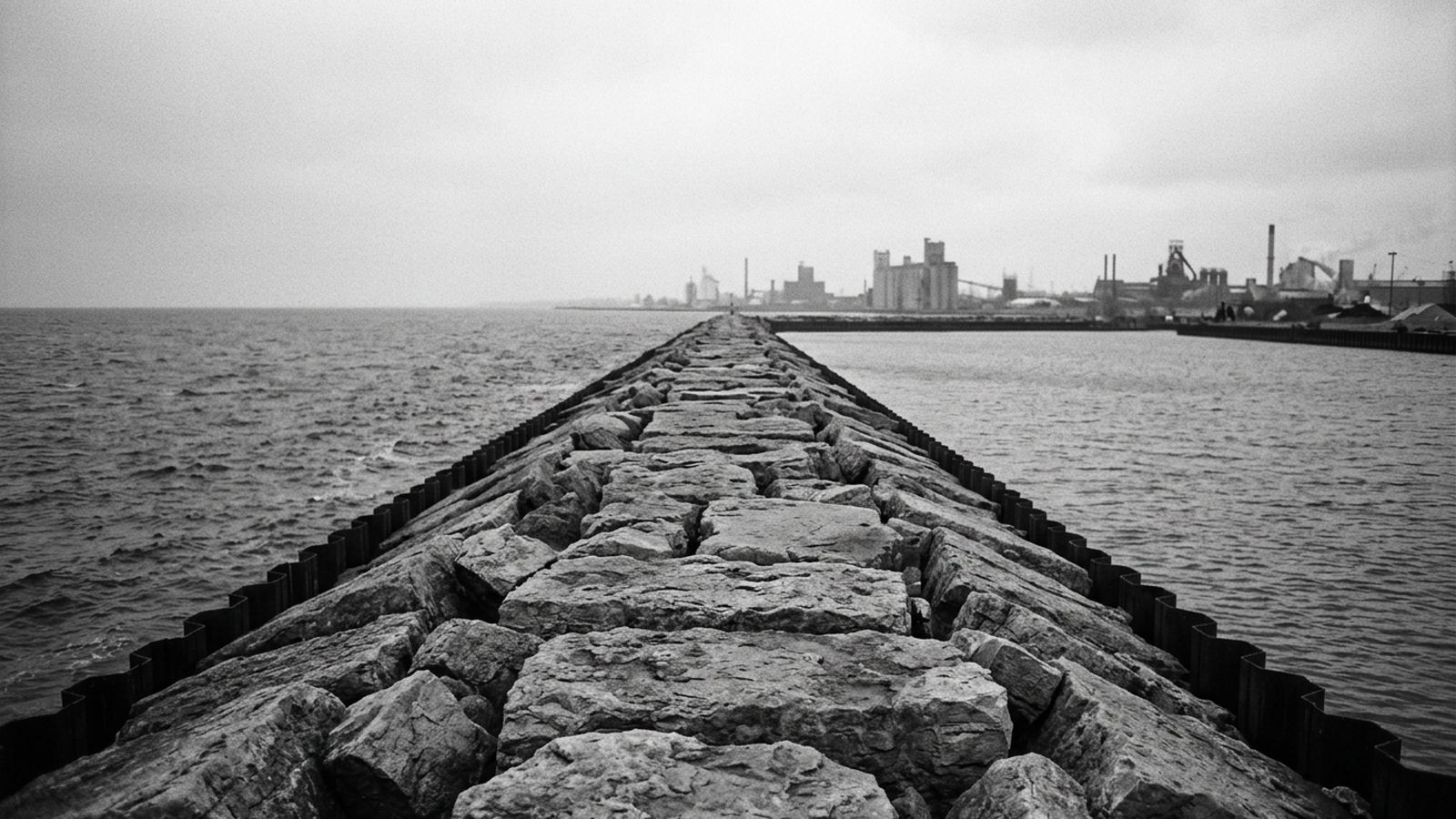 Looking along the top of a long stone breakwater wall at a harbor mouth, uneven capping stones and no handrail, open water on both sides, a distant industrial shoreline behind, flat overcast light with no people in frame