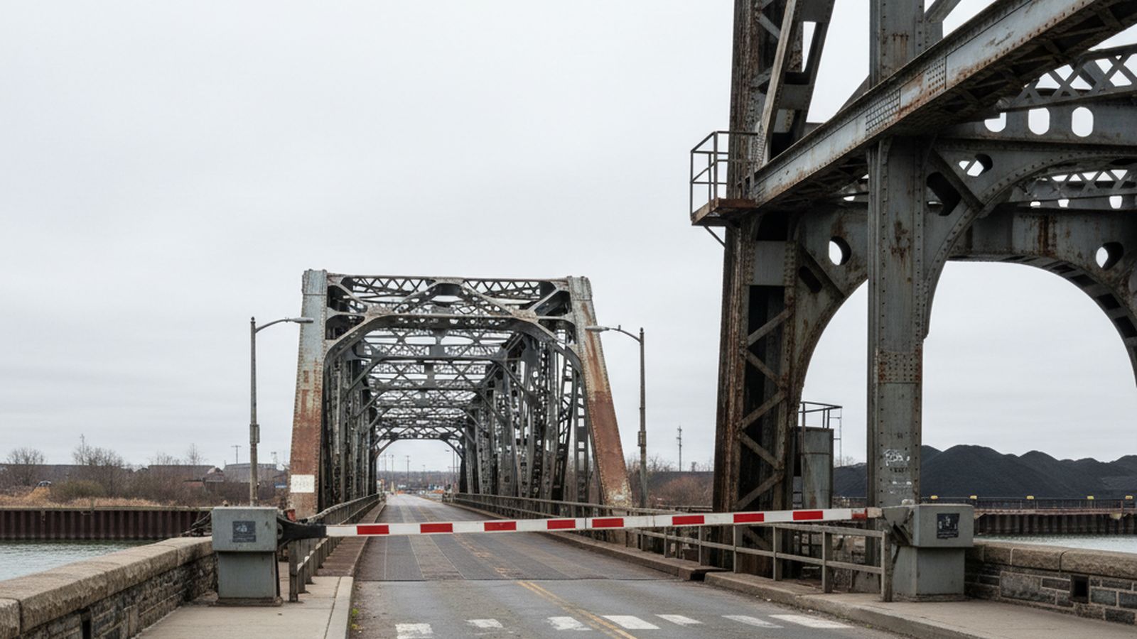A traffic gate lowered across the roadway of a movable bridge, seen from the fixed approach sidewalk, the movable deck beyond it and the steel tower leg rising at the right, worn road markings, flat overcast light, no vehicles and no legible signage
